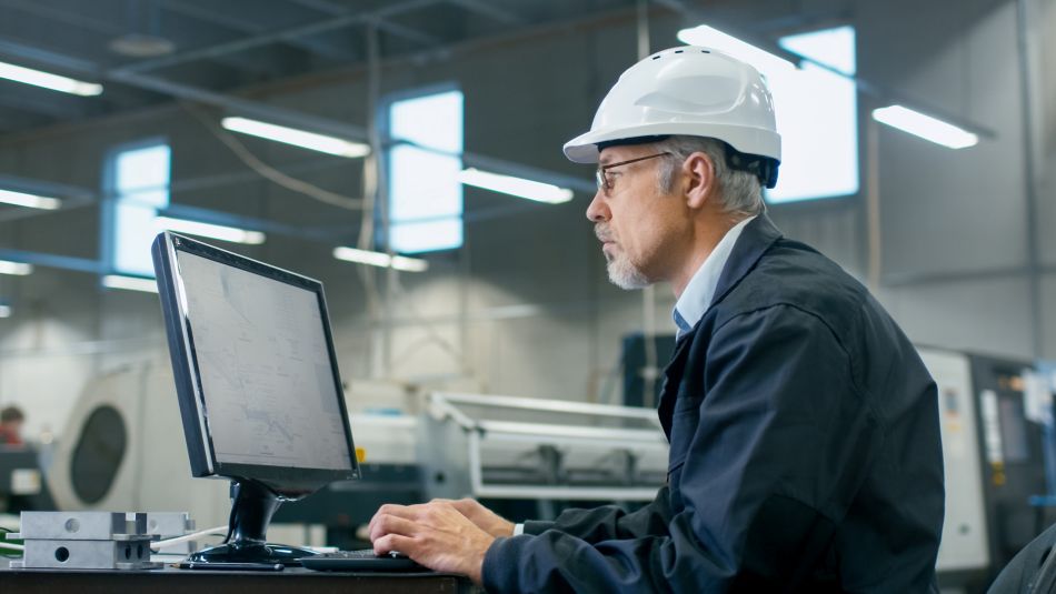 Senior engineer in glasses is working on a desktop computer in a factory.