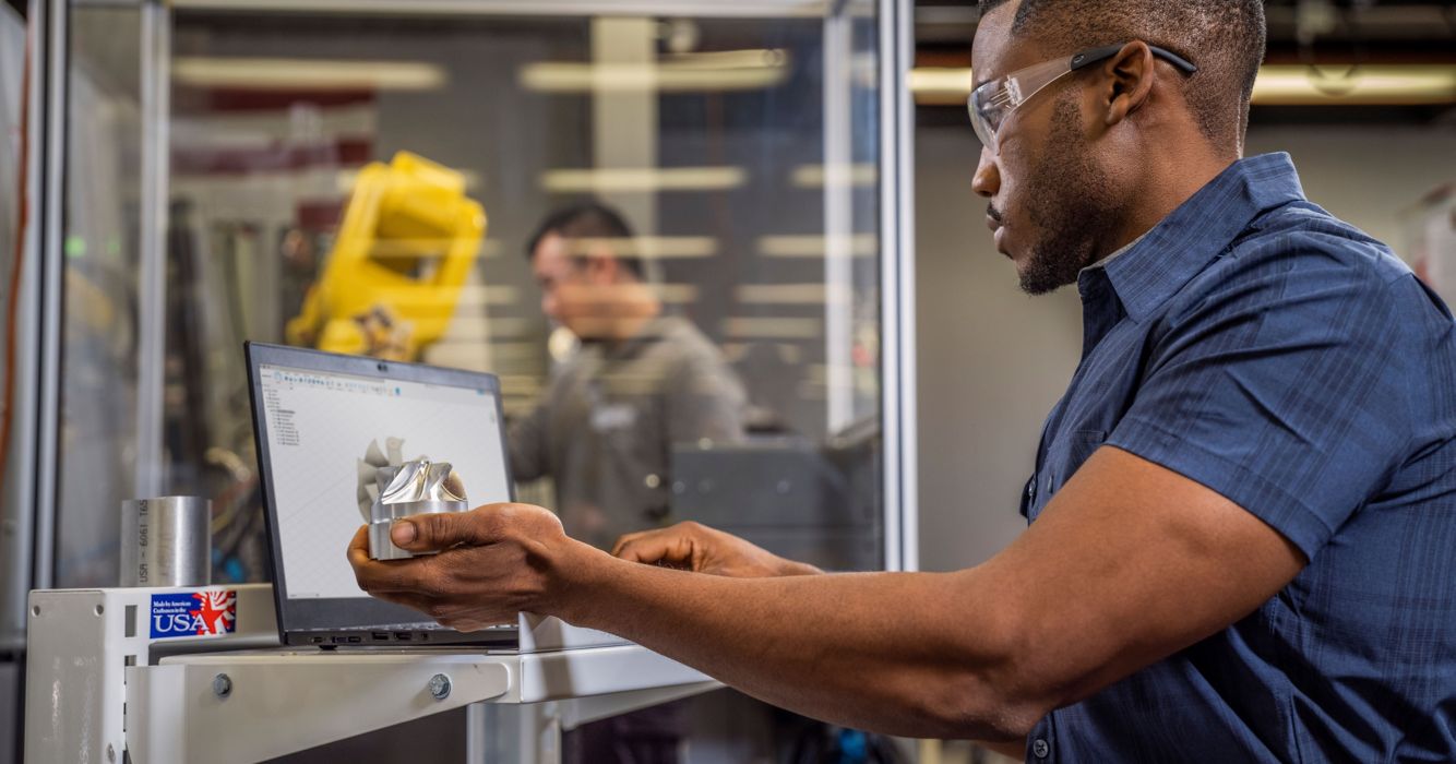 Man Looking at Machined Part and Working on a Computer