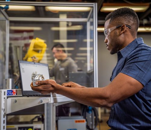 Man Looking at Machined Part and Working on a Computer